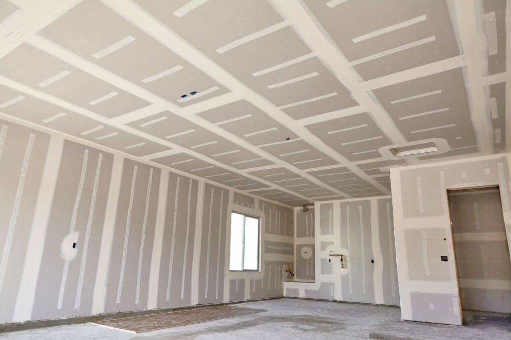 A newly built room with unfinished drywall on the walls and ceiling awaits the touch of skilled carpentry. The walls are covered with white joint compound at the seams, complementing a window on one wall. Construction materials for partitioning lie neatly organized on the floor.