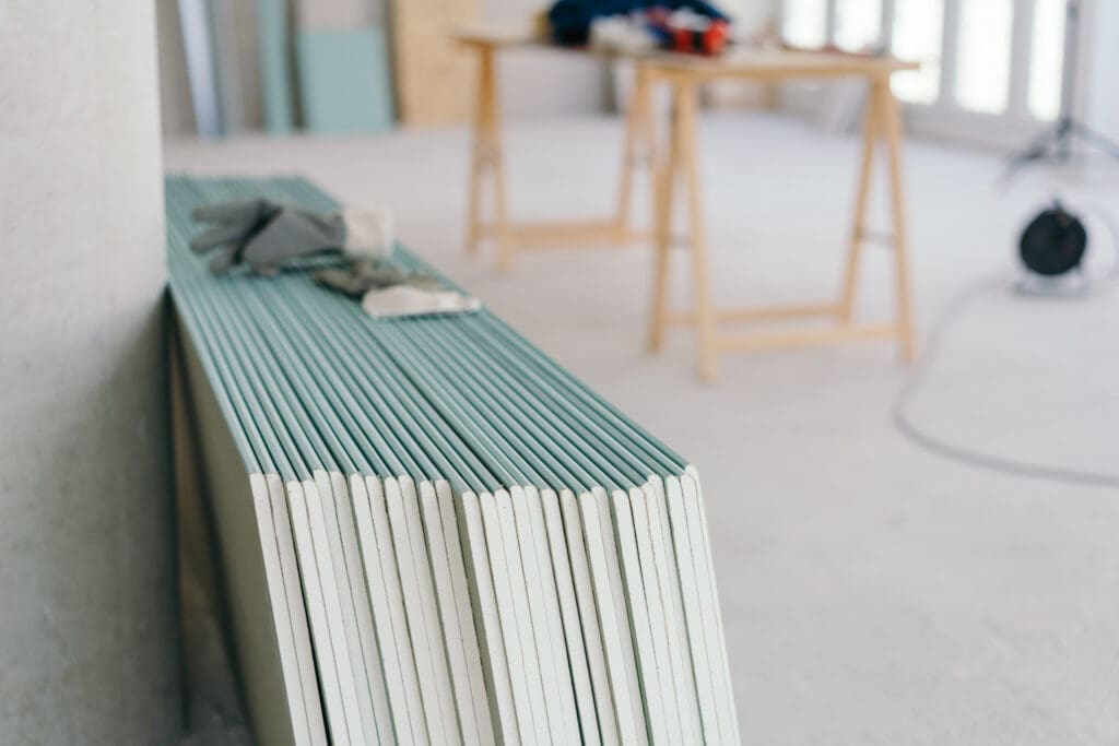 A stack of drywall panels leans against a wall in a bright, unfinished room, ready for the dry lining contractor. A pair of work gloves rests on top, with blurred work tables, tools, and equipment hinting at an office fit-out in progress in the background.
