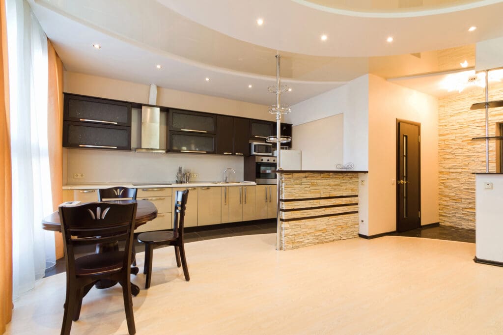Modern kitchen and dining area featuring a sleek design. Expert carpentry is evident in the dark upper cabinets, light lower cabinets, and stainless steel appliances. A round dining table with dark chairs sits near large windows, and the floor is light wood.
