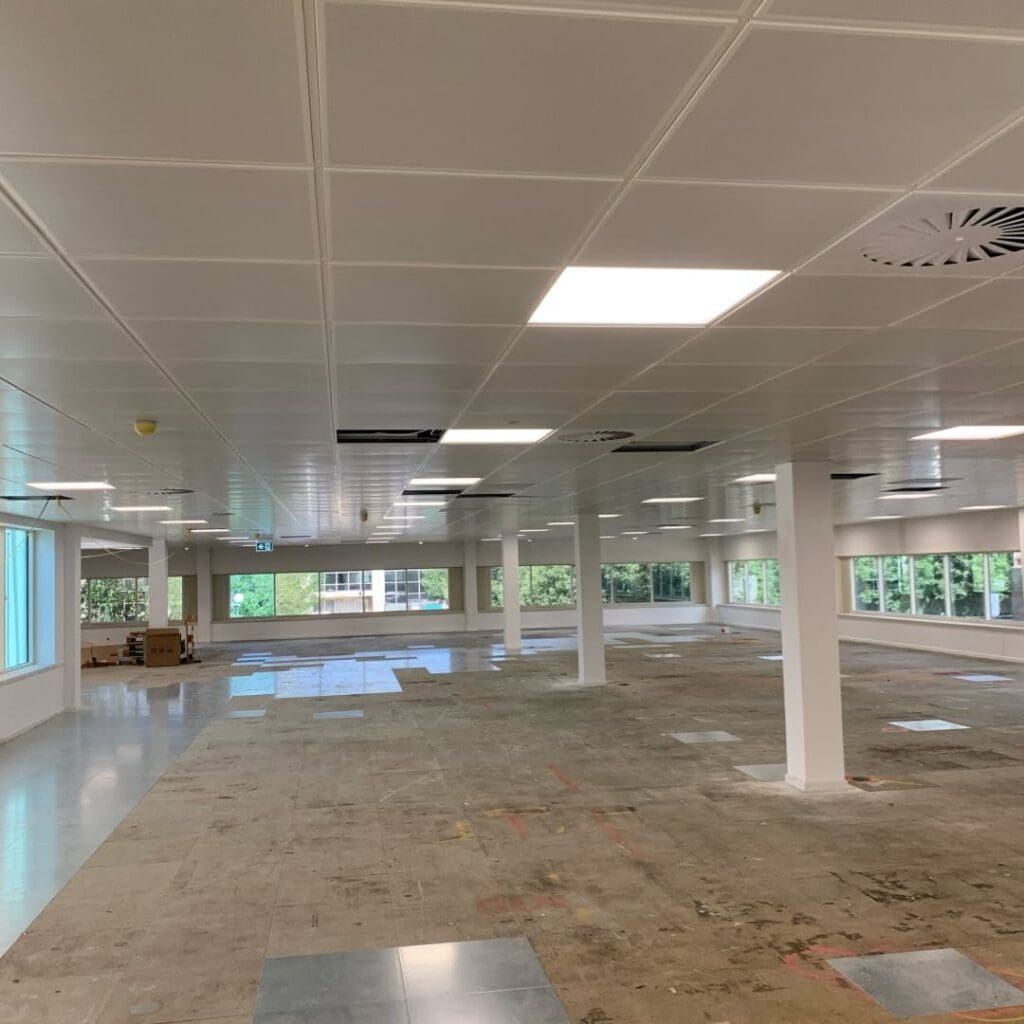 An empty, spacious office features suspended ceilings and support columns. Large windows flood the space with natural light. The floor is partially tiled, with some areas unfinished. A few cardboard boxes are stacked in the corner near the windows, awaiting carpentry work.