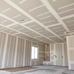 A room under construction with unfinished drywall on the walls and ceiling, featuring visible joints and seams, a window, open doorway, and exposed utility boxes. The floor appears bare and unfinished.
