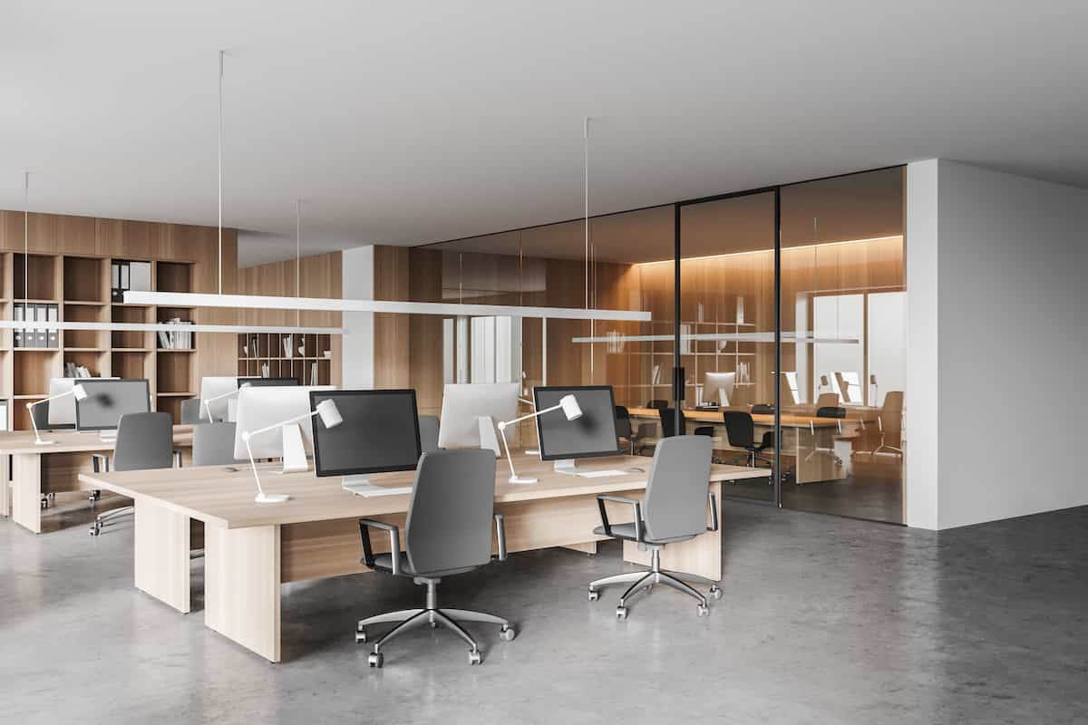 Modern open-plan office with wooden desks, gray chairs, desktop computers, and hanging lights. In the background, there is a glass-walled meeting room with additional chairs and a wooden accent wall.