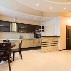 Modern open-plan kitchen with dark upper cabinets, light lower cabinets, stainless steel appliances, a small breakfast bar with stone accents, round dining table with chairs, and light wood flooring.