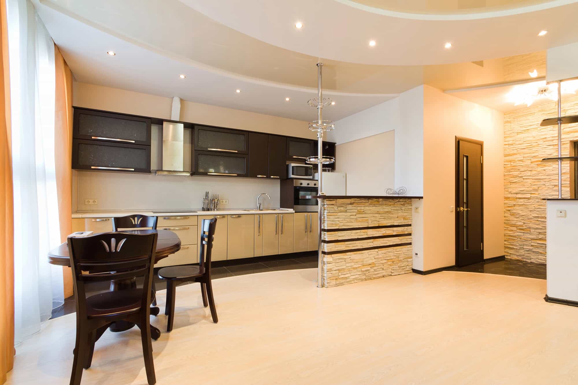 Modern open-plan kitchen with dark upper cabinets, light lower cabinets, stainless steel appliances, a small breakfast bar with stone accents, round dining table with chairs, and light wood flooring.